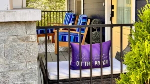 Front porch with a cushioned bench and blue-striped chairs, black railing in the foreground, leafy trees surrounding the space, and a yellow front door