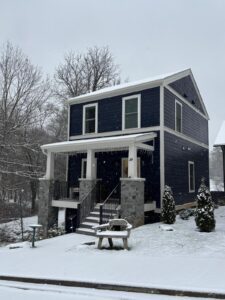 Cozy two-story home with a covered front porch, surrounded by trees and blanketed in fresh snow—perfect for a peaceful, inviting retreat.