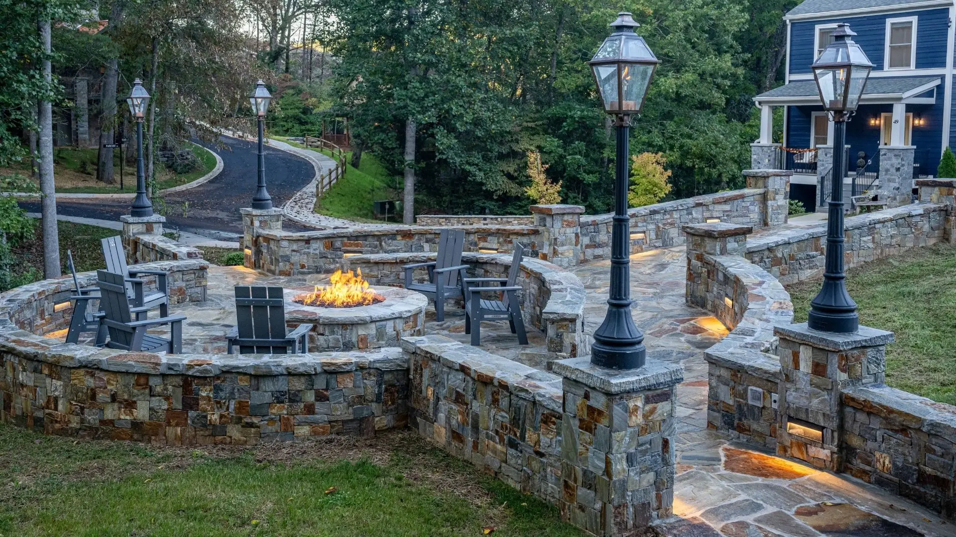 Daylight view of a stone gas firepit seating area with chairs, walkway, and nearby home in a mountain community in Franklin, NC.