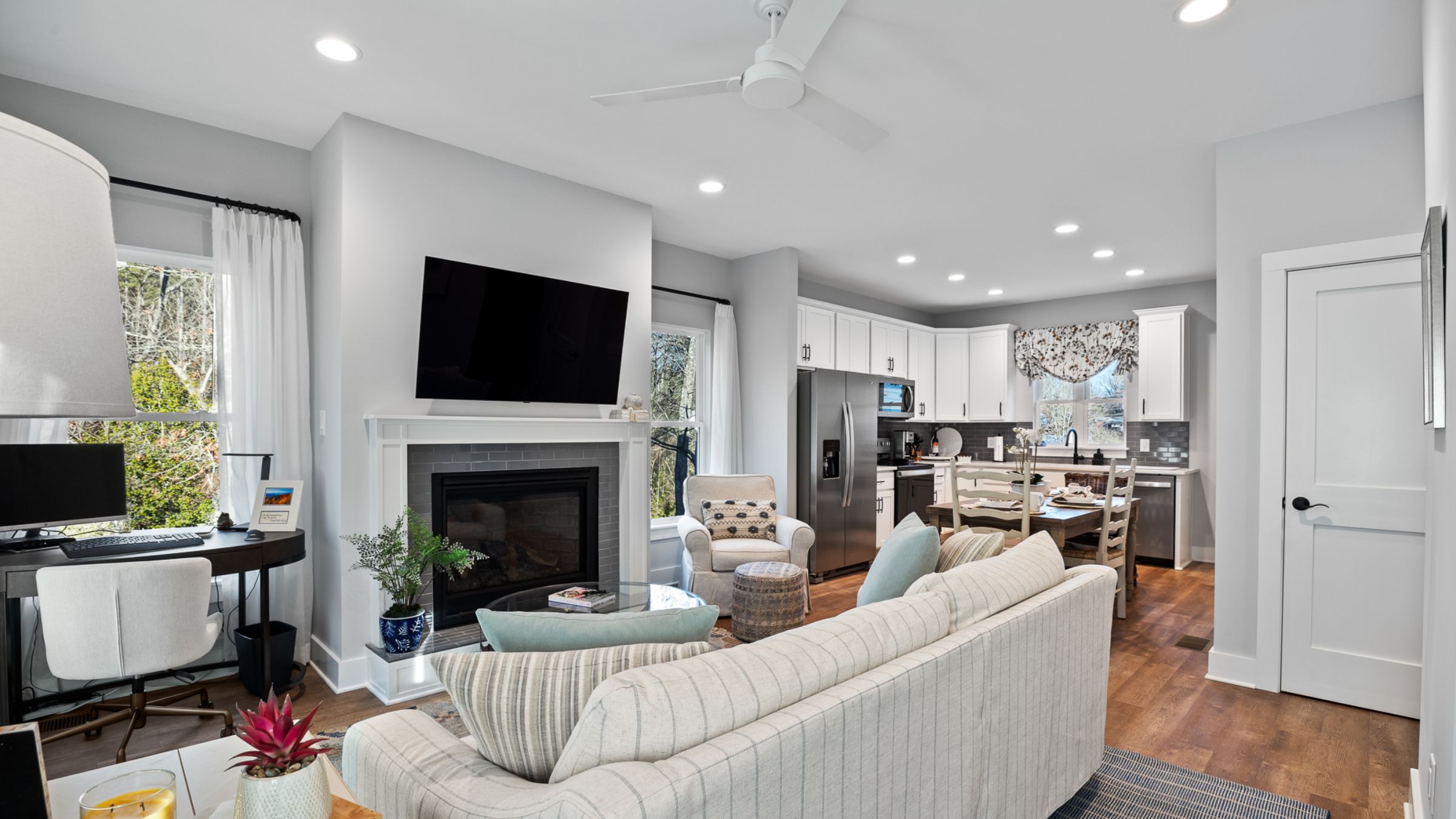 Open living room, dining room, kitchen fully furnished with wall mounted TV above fireplace. The foreground shows the living room and you can see dining area and kitchen in the background.